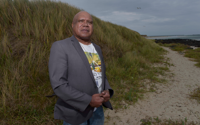 Archie Roach is wearing a white t-shirt and grey suit jacket standing in front of a grassy beachside hill